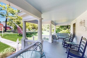 A covered porch with white columns, outdoor seating, and a view of a lawn and flag. - Classic village home with large porch, washer/dryer, cable, grill & partial AC (Manchester)