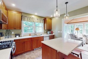 A kitchen with wooden cabinets, a central island, a stove, and a window with curtains. - Classic village home with large porch, washer/dryer, cable, grill & partial AC (Manchester)
