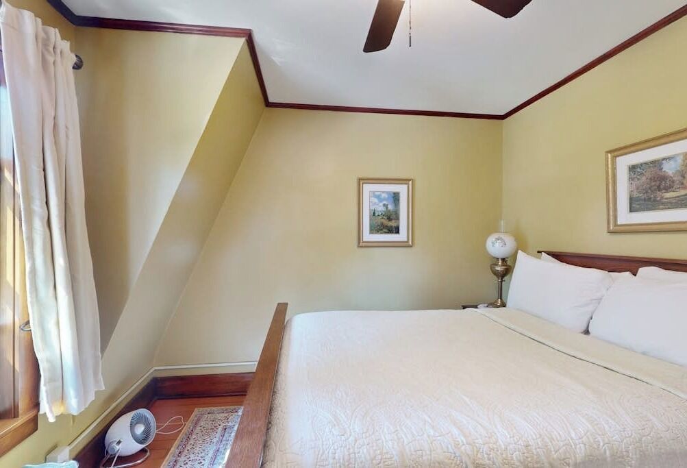 Une chambre à coucher comprenant un lit, un ventilateur de plafond, une table de chevet, une lampe, un tableau encadré et une fenêtre avec des rideaux.