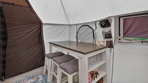 Een tent met een klein tafeltje en twee krukken, een plank met glazen, en een slaapgedeelte met een paars gordijn.