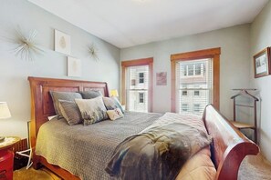 Ein Schlafzimmer mit einem hölzernen Bett, zwei Fenstern mit Jalousien, einem Nachttisch und Wanddekorationen.