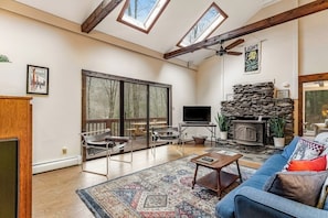 A spacious living room with a fireplace, large windows, and a ceiling fan. - Dog-friendly cabin with forest setting, woodstove & deck - near Killington (Plymouth)