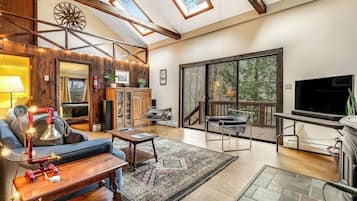 A spacious living area with a high ceiling, skylights, a fireplace, a TV, and a large sliding glass door to a deck.