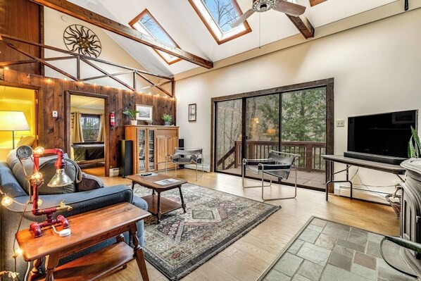 A spacious living area with a high ceiling, skylights, a fireplace, a TV, and a large sliding glass door to a deck. - Dog-friendly cabin with forest setting, woodstove & deck - near Killington (Plymouth)