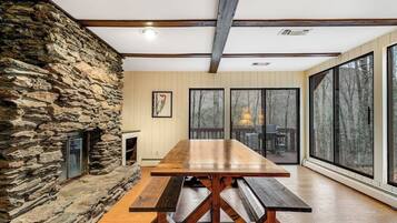 A stone fireplace, a wooden dining table with benches, and large windows with a view of trees.