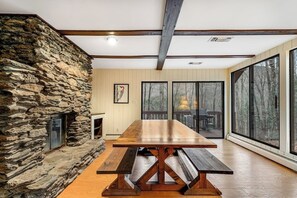 A stone fireplace, a wooden dining table with benches, and large windows with a view of trees. - Dog-friendly cabin with forest setting, woodstove & deck - near Killington (Plymouth)