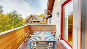 A balcony with a table and chairs, a view of a house, and a red-framed window. - Two-story townhome with forest/mountain view, gas fireplace, pool, gym & saunas (Proctorsville)