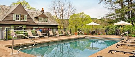 A swimming pool with lounge chairs and umbrellas, surrounded by a fence and trees.