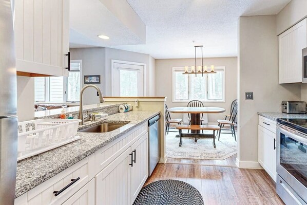 A modern kitchen with white cabinetry, granite countertops, a dining area with a round table and chairs, and a large window providing natural light. - Mountain-view condo with private balcony, fireplace, & updated interior (Stratton)