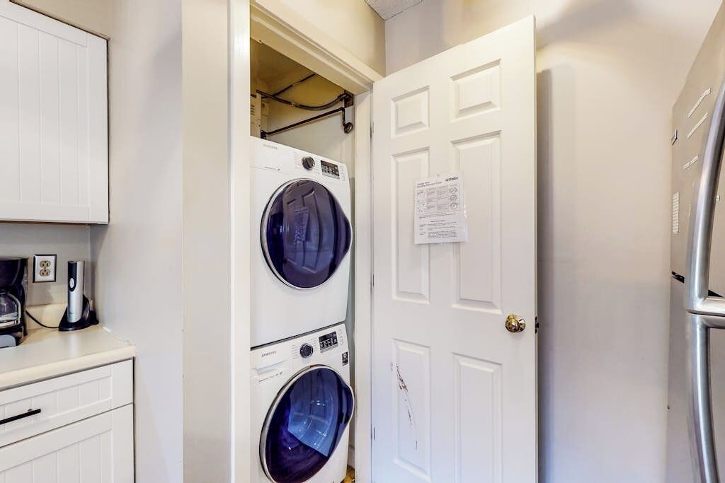 A laundry room with two stacked front-loading washing machines, a white door with a notice, and a refrigerator to the right.