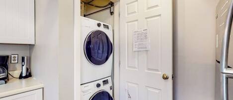 A laundry room with two stacked front-loading washing machines, a white door with a notice, and a refrigerator to the right.