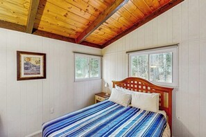 Ein Schlafzimmer mit Holzbalkendecke, einem Bett mit gestreifter Bettdecke, zwei Fenstern und einem gerahmten Bild an der Wand.