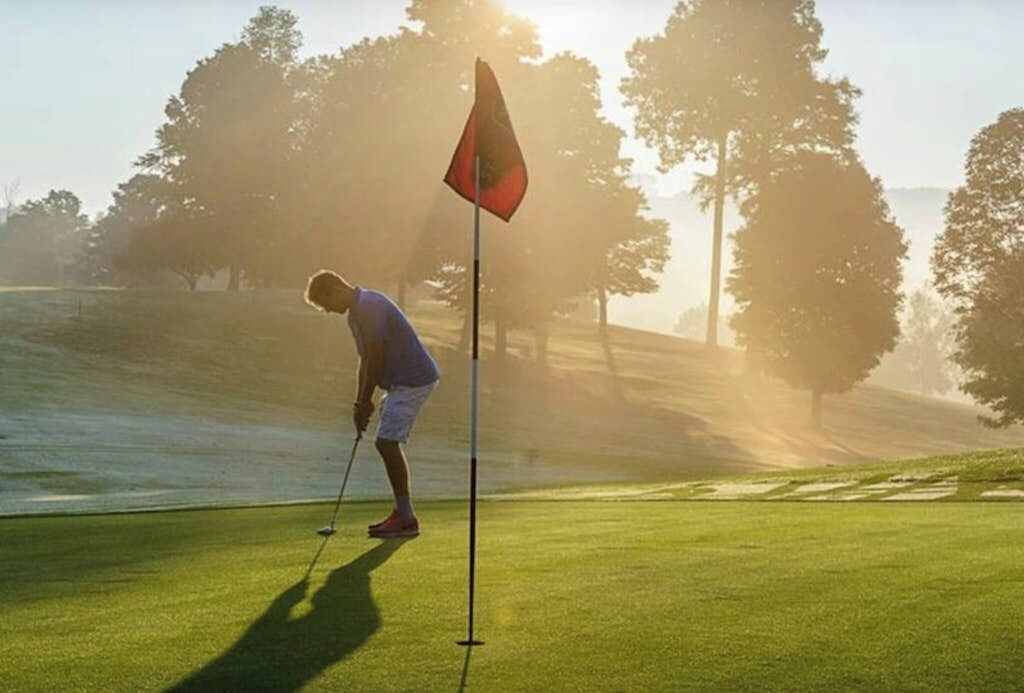 A person on a golf course with a flagstick, trees, and a clear sky.
