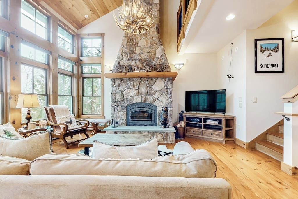 A spacious living room with a stone fireplace, large windows, a television, and a cozy seating area.