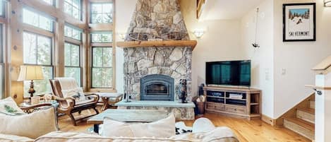 A spacious living room with a stone fireplace, large windows, a television, and a cozy seating area.