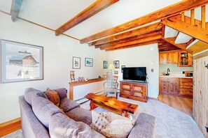 Ein Wohnzimmer mit einer Couch, einem Holztisch und einem Fernseher. Es gibt eine Küchenzeile mit Holzschränken und einem Esstisch.