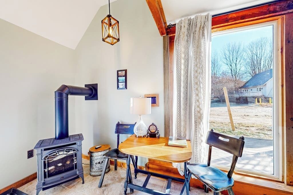 Una habitación acogedora con una estufa de leña, una mesa de comedor, sillas, una lámpara y un ventanal con vistas al exterior.