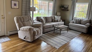 A living room with a sofa, armchair, and coffee table, large windows with curtains, and a hanging light fixture. -  3-bedroom house in tranquil Millinocket (Millinocket)