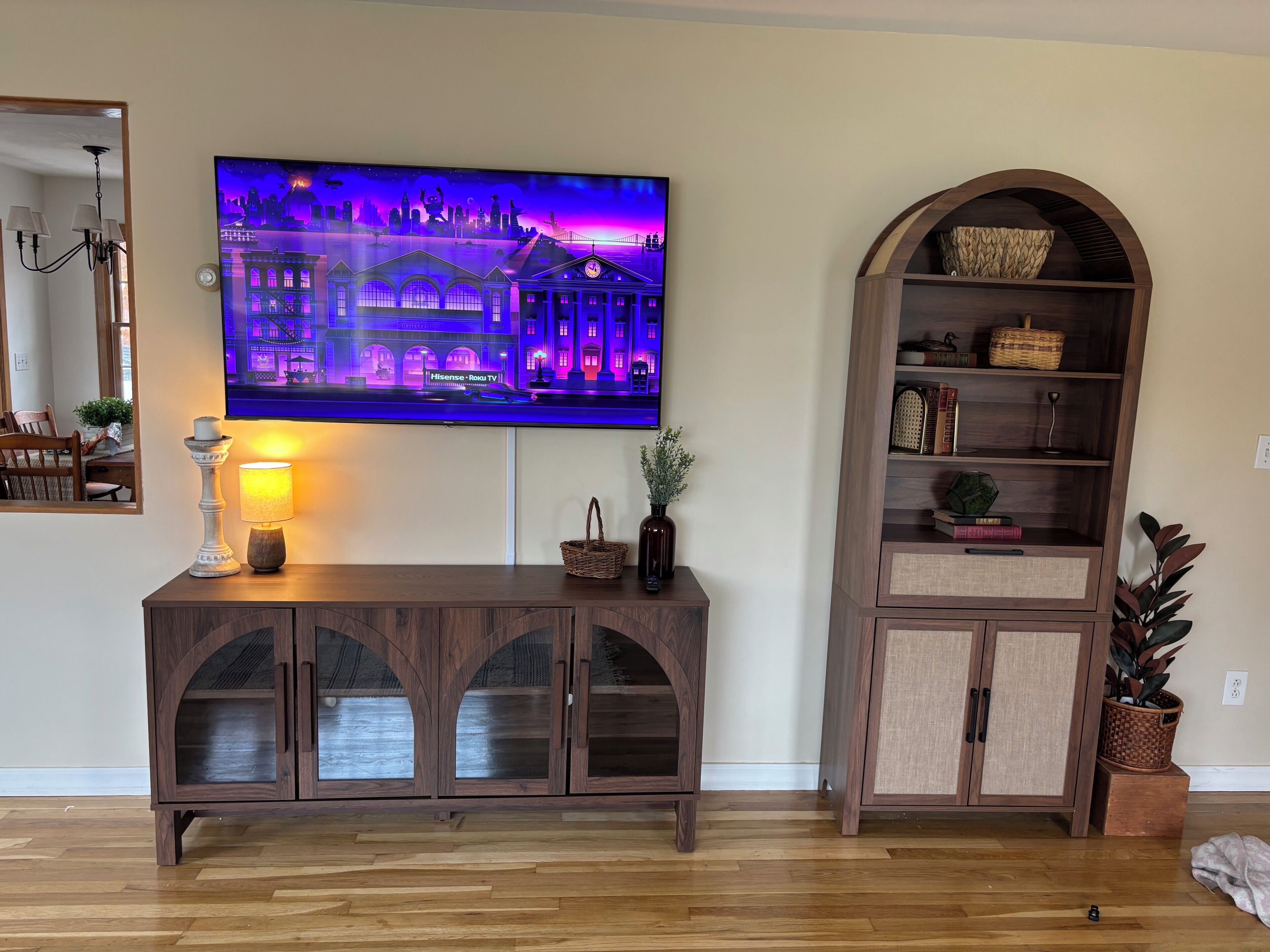 Ett vardagsrum med en tv-skärm som visar en stadsbild, ett träskåp med glasdörrar, en lampa och en bokhylla.