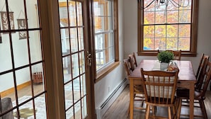 A dining area with a wooden table and chairs, a chandelier, and a view through large windows. -  3-bedroom house in tranquil Millinocket (Millinocket)