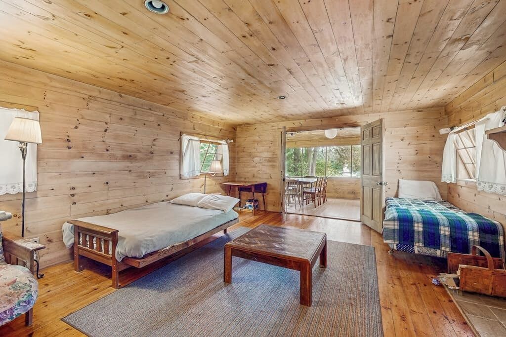 A wooden cabin with two beds, a dining table, and a view through a large window.