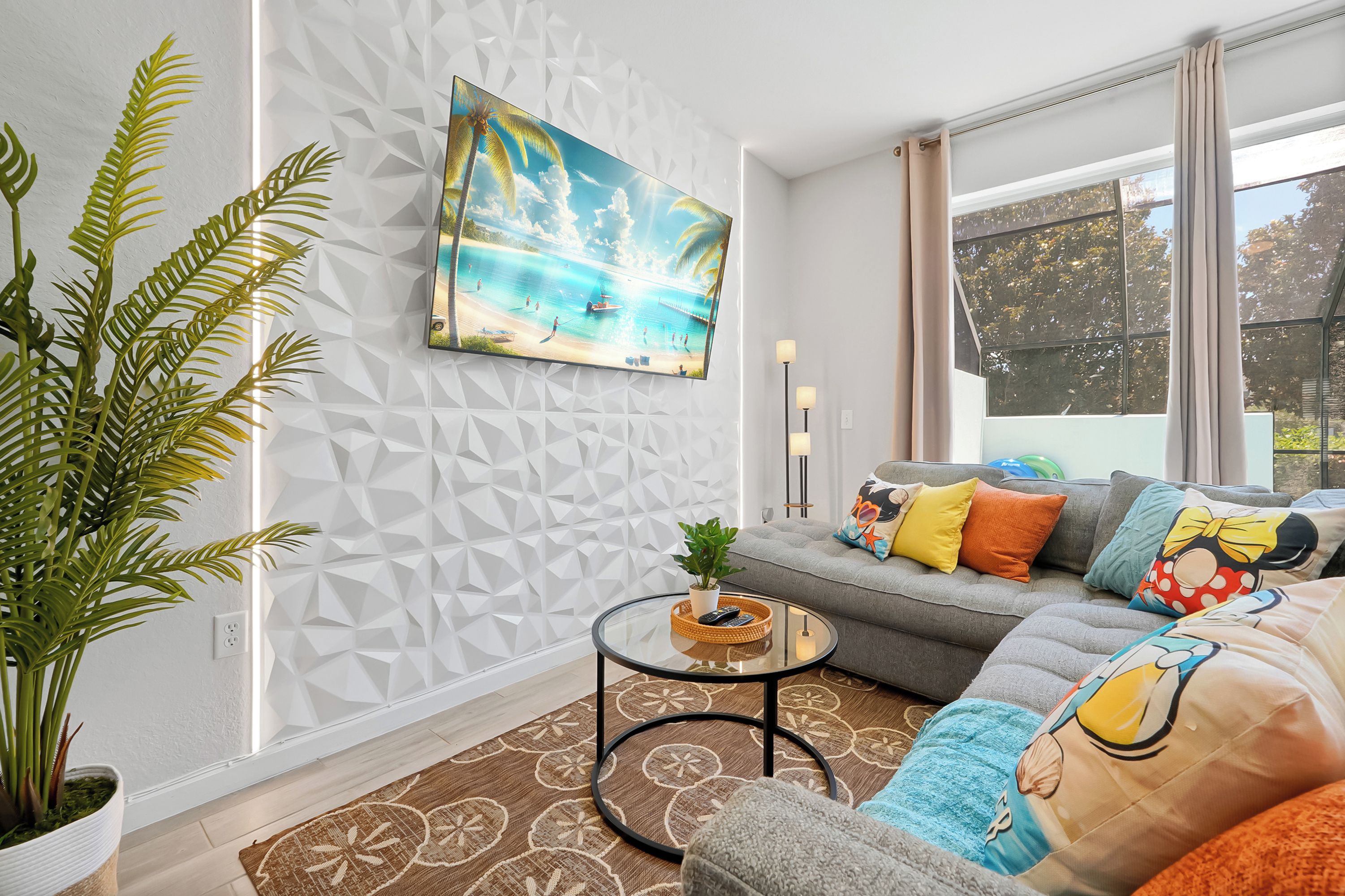 Un salon moderne avec un motif géométrique sur un mur, une grande peinture sur le thème de la plage, un canapé agrémenté de coussins colorés, une table basse en verre avec une plante, et une vue dégagée sur l’extérieur grâce à une grande fenêtre.