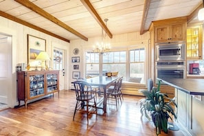 Ein Zimmer mit Holzwänden, einem Esstisch, Stühlen, einer Chandelierleuchtre und einer Küche mit Mikrowelle und Backofen.