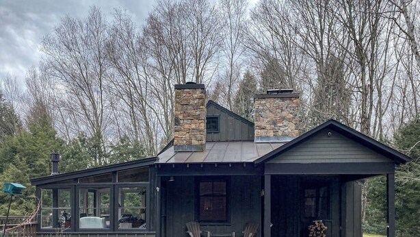 A cabin with a stone chimney, a porch with chairs, and a view of trees.
