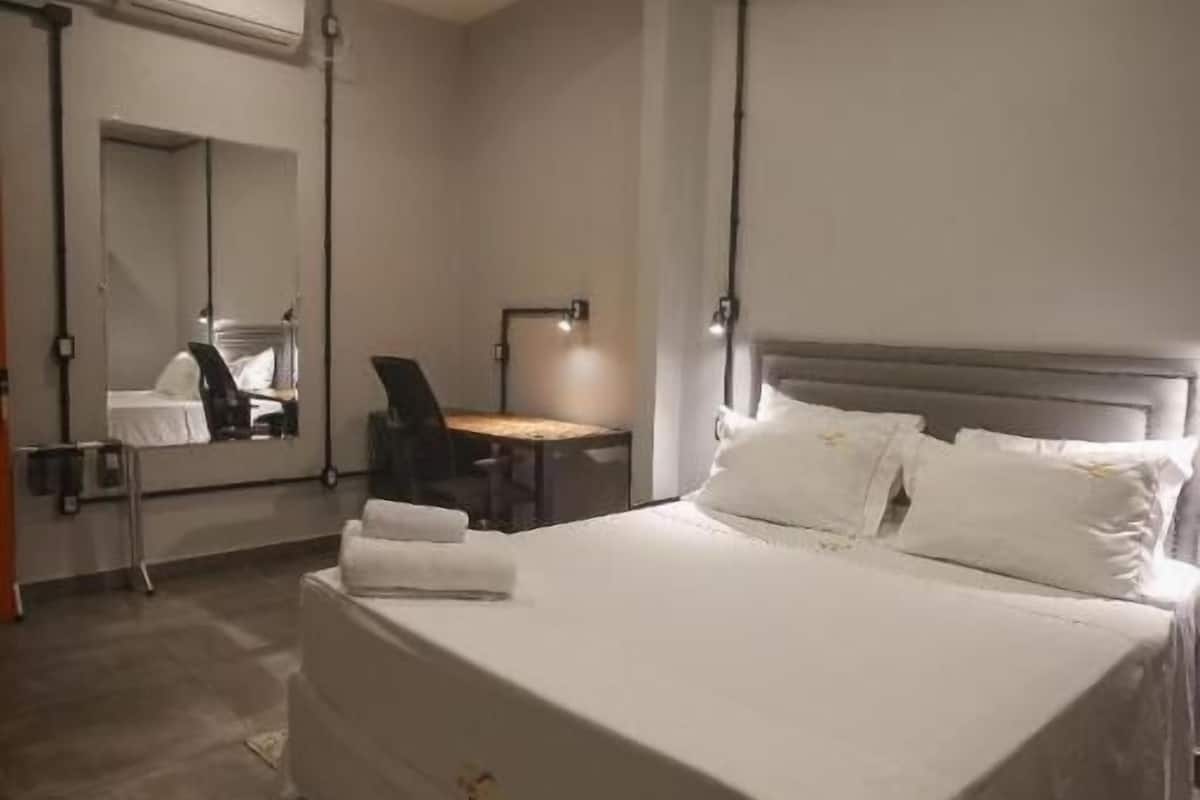 A hotel room with a large bed, a desk, a chair, a mirror, and a wall-mounted air conditioning unit.
