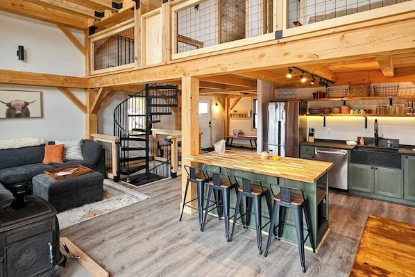 A spacious loft-style living area with a central wooden bar, high ceilings, exposed beams, a spiral staircase, and a kitchen with green cabinets.