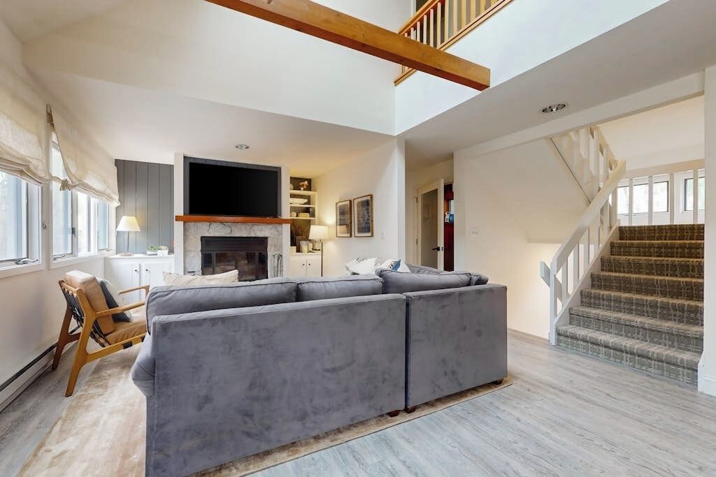 A spacious living area with a gray sectional sofa, a fireplace, a flat-screen TV, and a staircase.