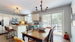 A modern kitchen with white cabinets, a dining table with chairs, a chandelier, and a large window. - Delightful house w/ hot tub, wrap-around deck, ping-pong-pool table, fireplaces (Ludlow)
