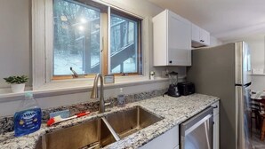 A kitchen with a stainless steel sink, a window with a view of a snowy deck, white cabinets, and a refrigerator.