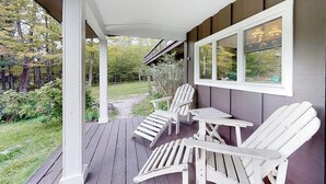 Varanda coberta com cadeiras Adirondack e uma mesa, com vista para um jardim e uma área arborizada.