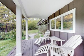 Eine überdachte Veranda mit Adirondack-Stühlen und einem Tisch, mit Blick auf einen Garten und einen bewaldeten Bereich.