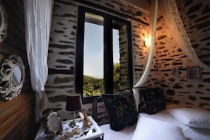 A room with a stone wall, a bed with white linens, a window with a view, and a lamp on a bedside table. - Filoxenia-Drakeia (Drakia)