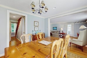 A dining area with a wooden table and chairs, a chandelier, and a view into another room with a sofa and TV. - Spacious village house, dog-friendly, patio, fireplace, a/c, air hockey, w/d (Manchester)