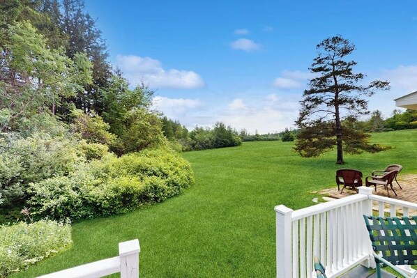 A well-maintained lawn with a white fence, a wooden deck with chairs, and a large tree.