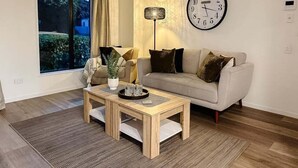 A living room with a sofa, a coffee table, a clock, and a window with curtains. - Stadium Gatehouse - Bookahome (Central City Christchurch)