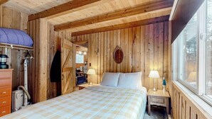 Ein Schlafzimmer einer Holzhütte mit einem Bett, zwei Nachttischen, einem Fenster und einer Tür zu einem anderen Raum.