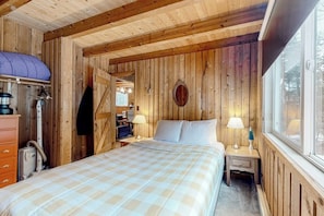 Un dormitorio de cabaña de madera con una cama, dos mesitas de noche, una ventana y una puerta que da a otra habitación.