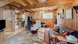 Ein rustikales Interieur einer Holzhütte mit Holzbalkendecke, einer Couch, einem Fernseher, einem Essbereich und einer durch eine offene Tür sichtbaren Küche.