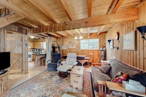 Interior rústico de una cabaña con techo de madera, un sofá, un televisor, un comedor y una cocina visible a través de una puerta abierta.