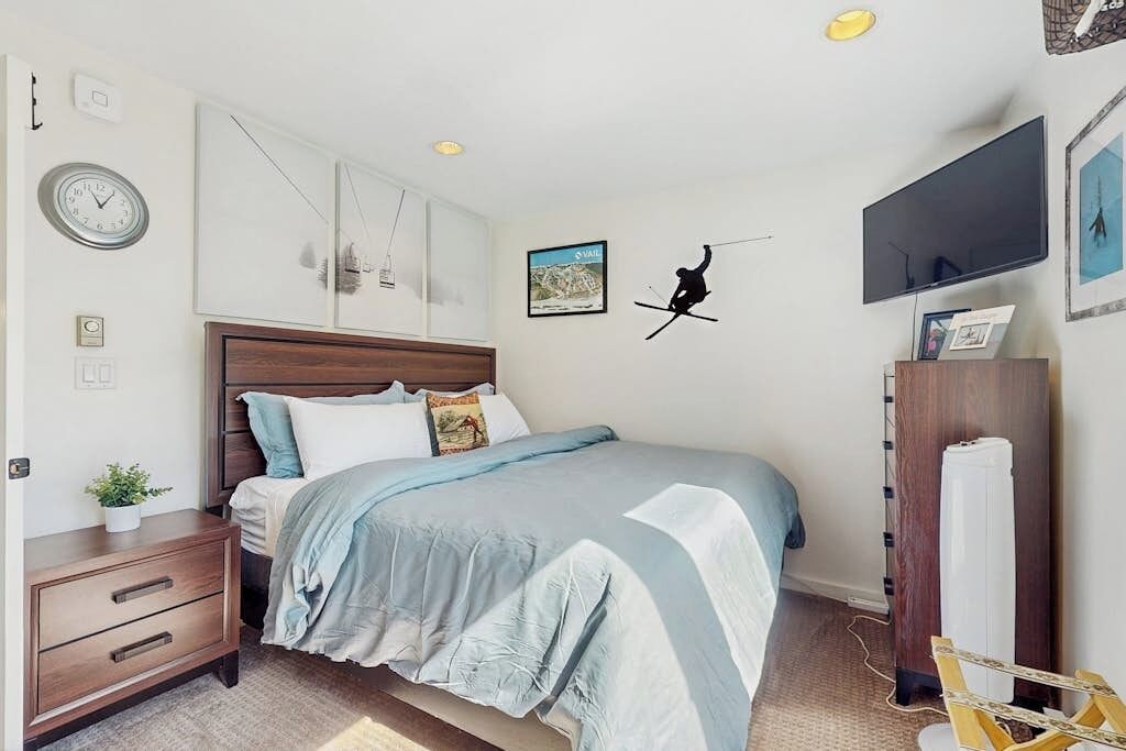 Un dormitorio con cama, mesita de noche, reloj de pared, cuadros enmarcados y televisión.