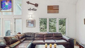 Ein Wohnzimmer mit einer großen, ledernen L-Form-Couch, einem Couchtisch mit Kerzen und einem großen Fenster mit Blick auf Bäume.