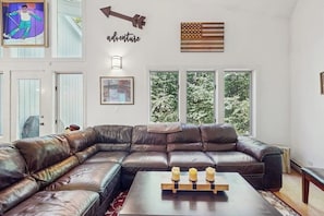 A living room with a large L-shaped leather sofa, a coffee table with candles, and a large window with a view of trees. - Ski-in/ski-out townhouse w/ trail access, fireplace, balcony, pool, hot tub (Stratton)