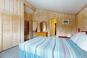 Ein Zimmer mit Holzwandverkleidung, einem Bett mit gestreifter Bettwäsche, einer Holzkommode, einem Spiegel und einer Tür zu einem weiteren Raum.