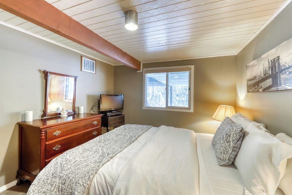 Un dormitorio con cama, cómoda, televisor, ventana y una obra de arte en la pared.