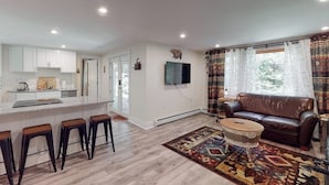 Una sala de estar moderna con un sofá de cuero marrón, un tapete estampado y una zona de cocina con armarios blancos y un taburete de bar.