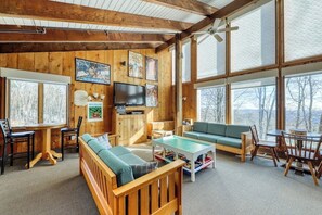 Interior acogedor de una cabaña con paredes de madera, un televisor grande y vistas al exterior.